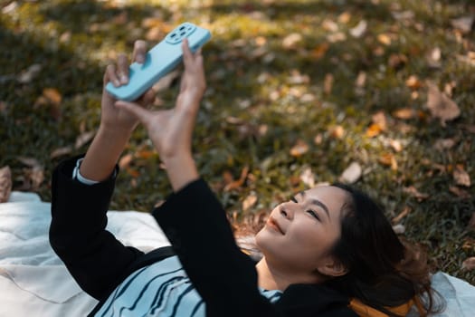 Carefree beautiful businesswoman lying on grass and using mobile phone at garden or park in autumn.