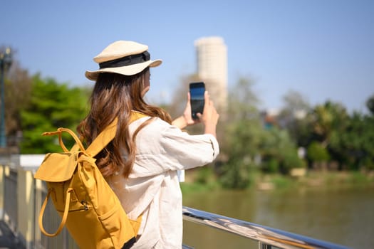 Back view of tourist woman with backpack taking photo with smartphone on a bridge over a canal.