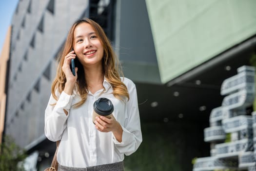 Beautiful woman walking outside in street and calling talking on mobile phone outdoors near office, portrait lifestyle businesswoman with bag cell to customer while holding coffee paper cup takeaway