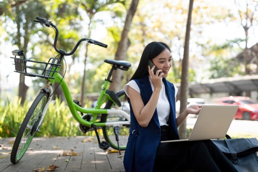 Asian businesswoman with bicycle using laptop and smartphone and sitting outside the office building. Woman commuting on bike go to work. Eco friendly vehicle, sustainable lifestyle concept.