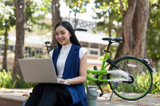 Asian businesswoman with bicycle using laptop and sitting outside the office building. Woman commuting on bike go to work. Eco friendly vehicle, sustainable lifestyle concept.