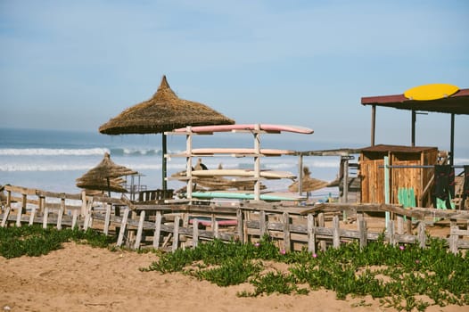 Nature background of a tropical sandy beach with stacked surfing boards and straw umbrellas, parasols against background of Atlantic ocean with beautiful waves on sunny day. Summer holidays concept