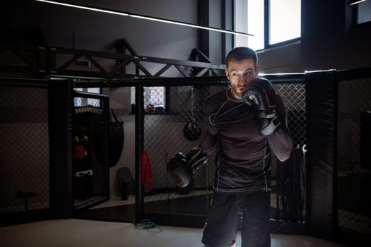Focused expressive male MMA fighter in boxing gloves training punches inside octagonal cage in gym setting, practicing shadow fighting alone