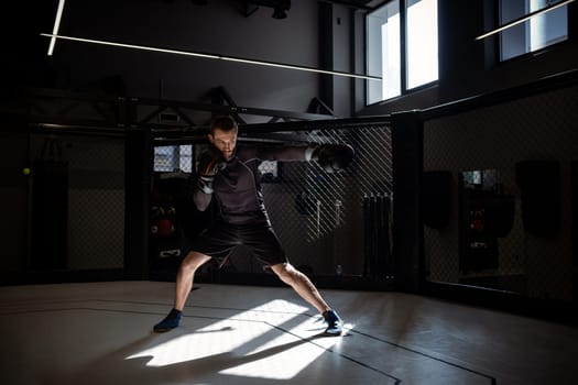 Determined concentrated young adult fighter immersed in shadowboxing, perfecting precise punches and footwork in rays of sunlight in boxing cage in modern urban gym setting