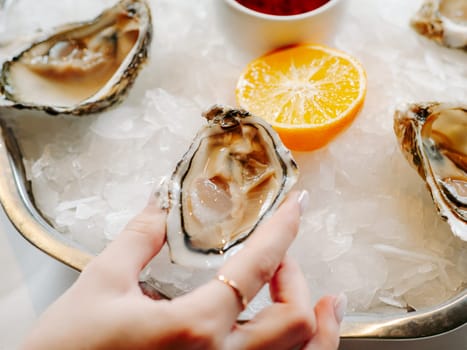 Woman having oysters at table. Fresh oysters on large plate with ice and lemon. Healthy food, gourmet food, restaurant food. Mediterranean cuisine, girl eats, oysters on ice