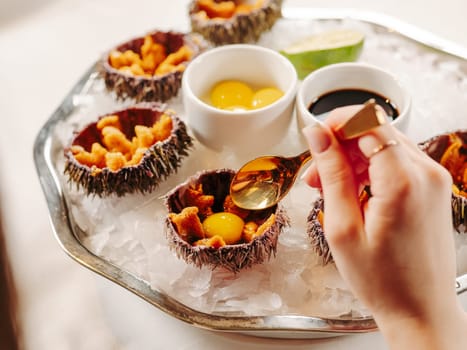 Woman having fresh sea urchins at table. Fresh sea urchins on large plate with ice and lime. Healthy food, gourmet food, restaurant food. Mediterranean cuisine, girl eats, sea urchins on ice