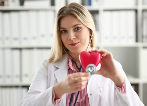 Beautiful smiling blond female doctor hold in arms red toy heart closeup. Cardio therapeutist student education CPR 911 life save physician make cardiac physical pulse rate measure arrhythmia