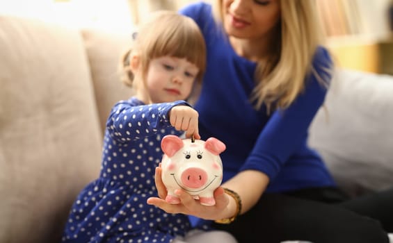 Child little girl arm putting pin money coins into happy pink faced piglet slot closeup. Making effective future needs savings collect dollar gift benefit present home leisure concept
