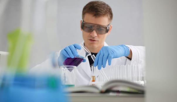 A male chemist holds test tube of glass in his hand overflows a liquid solution of potassium permanganate conducts an analysis reaction takes various versions of reagents using chemical manufacturing.