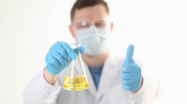 A portrait of a young surgeon chemist doctor holds thumbs up looks at a container with a yellow liquid and a mask is fought with viruses and a vaccine for vaccines against diseases.