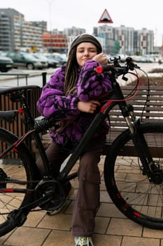 A pretty young woman with piercings and dreadlocks dressed in a purple fur coat rides a bicycle.