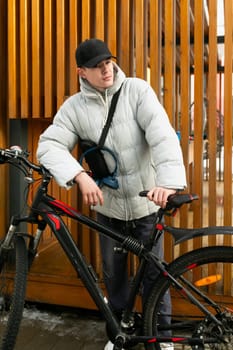 A young Caucasian man with a gray jacket and black cap walks around the city with a bicycle.