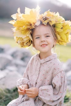 Little smiling girl in a wreath of autumn leaves sits on a stone on the lawn. High quality photo