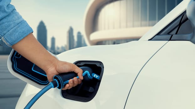 Asian man insert EV charger and recharge his electric car from future charging station with blurred background of cityscape. Smart and futuristic sustainable energy infrastructure. Peruse