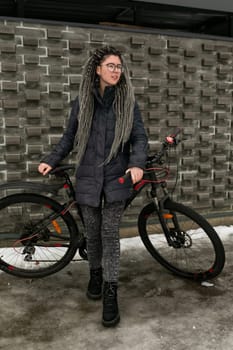Cute young woman with dreadlocks hairstyle dressed in a winter jacket rides a bicycle.