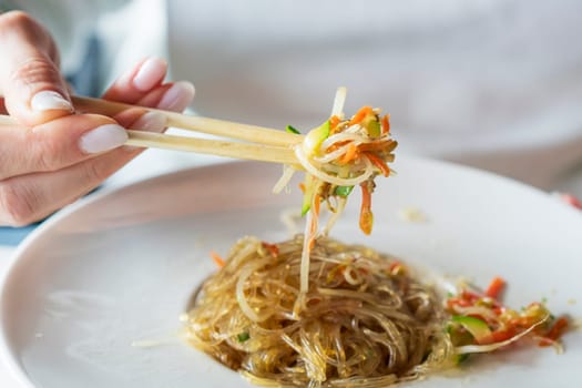 A woman with chopsticks in hand takes stir-fried glass noodles with vegetables, Japchae (also spelled chapchae or 잡채 in Korean) is a popular Korean dish.