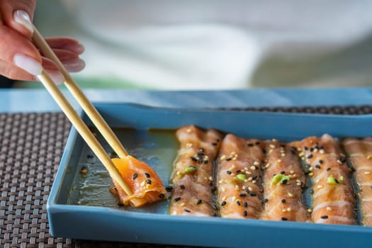 A woman with chopsticks in hand takes thin slices of salmon marinated with sesame, herbs and sauce. Healthy vegetarian food, raw salmon meat. Delicious Asian dish served on restaurant table