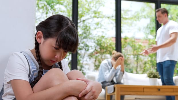 Stressed and unhappy young girl huddle in corner, cover her ears blocking sound of her parent arguing in background. Domestic violence at home and traumatic childhood develop to depression. Synchronos