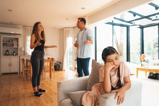 Annoyed and unhappy young girl sitting on sofa trapped in middle of tension by her parent argument in living room. Unhealthy domestic lifestyle and traumatic childhood develop to depression.Synchronos