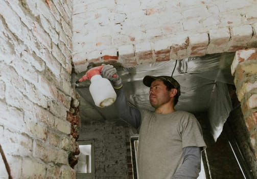 One young handsome Caucasian brunette man in casual clothes stands on a stepladder and sprays water from a spray bottle on bricks in a doorway under the ceiling in an old house, bottom side close-up view.