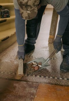 One young Caucasian man, unrecognizable in a gray casual work uniform, sweeps construction debris into a dustpan with a broom after laying bricks in a doorway, standing at an angle in an old ruined house, close-up side view.