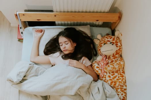 One beautiful little Caucasian brunette girl with flowing hair sleeps sweetly early in the morning in a wooden bed with a soft toy giraffe covered with a gray blanket near the radiator, close-up top view.