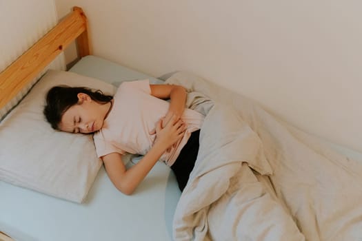 One beautiful little Caucasian brunette girl with a sad emotion on her face, loose long hair, sleeps in bed, curled up in pain and holding her stomach with both hands, close-up view from above.