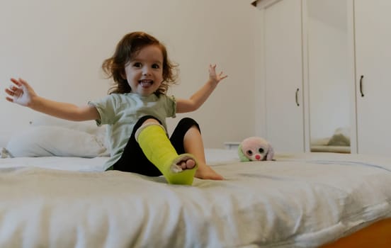 One beautiful little Caucasian girl with a green cast on her leg sits on the bed and cheerfully waves her arms with a happy smile on her face early in the morning in the room watching a cartoon on TV, side view close-up.