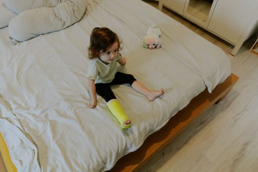 One beautiful little Caucasian baby girl with a green cast on her leg sits on the edge of the bed, watching a cartoon on TV in the morning in the room, top view close-up.
