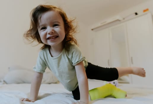One beautiful little Caucasian girl with a happy smile on her face, fooling around, stands on her knees with a green cast on the bed early in the morning in the room, close-up bottom view with depth of field.