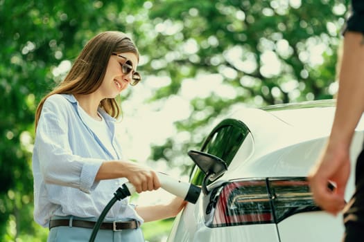 Lovely young couple wearing sun glasses recharging battery for electric car during road trip travel EV car in natural forest or national park. Eco friendly travel during vacation and holiday. Exalt