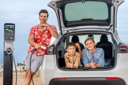 Family vacation trip traveling by the beach with electric car, lovely family sit on the trunk, charging EV car battery with green and sustainable energy. Family travel and eco-friendly car. Perpetual