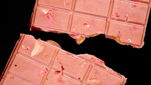 A bar of pink ruby chocolate with freeze-dried strawberries and almonds close-up, isolated black background. A healthy dessert based on berries and nuts