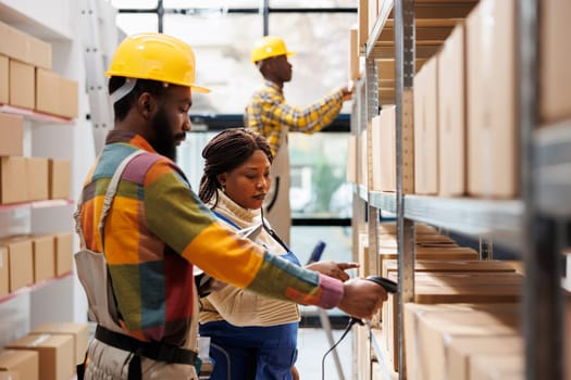 Warehouse manager supervising assistant scanning parcel barcode and pointing at cardboard box on shelf. African american storehouse worker using scanner on package and working with supervisor