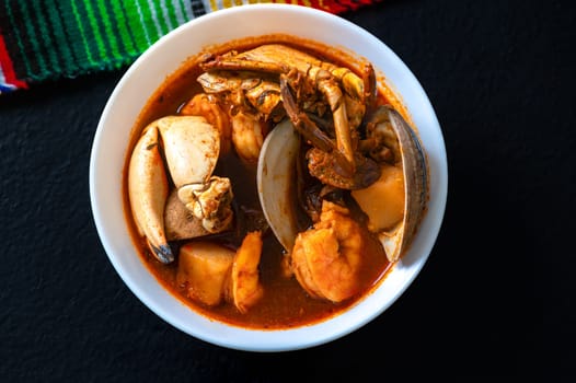 Mexican caldo de mariscos, a spicy and warming seafood stew made with chiles, fish, shrimp, crab and clams. Served in a white bowl on a black table. Flat lay.