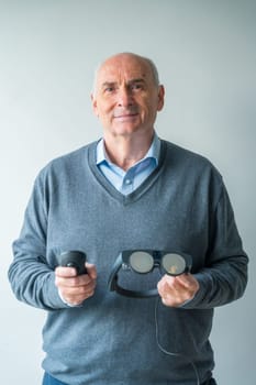 Vertical studio portrait with grey background of an aged man holding a mixed reality goggles and remote control