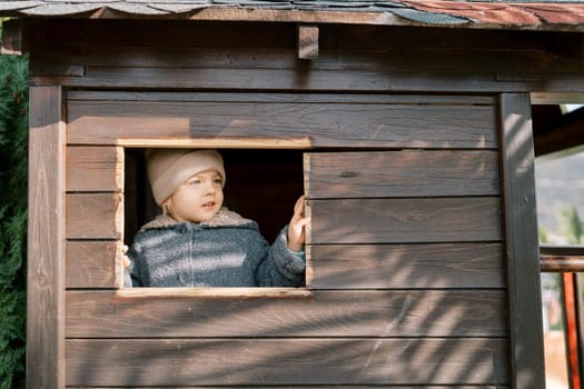 Little girl looks out of the window of a toy wooden house on the playground. High quality photo