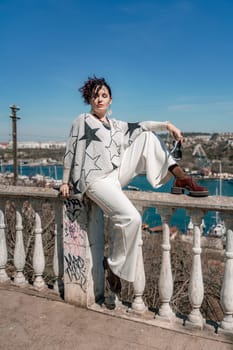 Woman walks around the city, lifestyle. A young beautiful woman in white trousers and a sweater sits on a white fence with balusters and overlooks the sea bay and the city