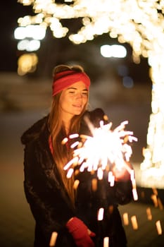 Woman holding sparkler night while celebrating Christmas outside. Dressed in a fur coat and a red headband. Blurred christmas decorations in the background. Selective focus.