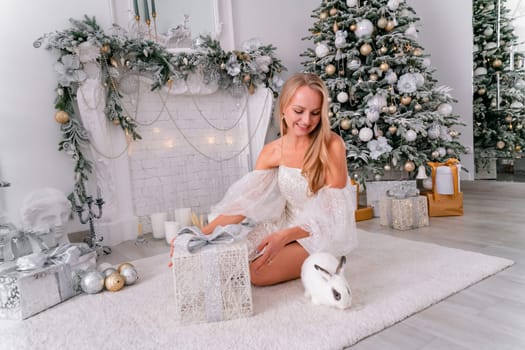 Woman holding a white rabbit symbol of the year 2023. Close-up of a beautiful young blonde woman holding a rabbit in a sparkly dress. She sits in a Christmas decorated room.