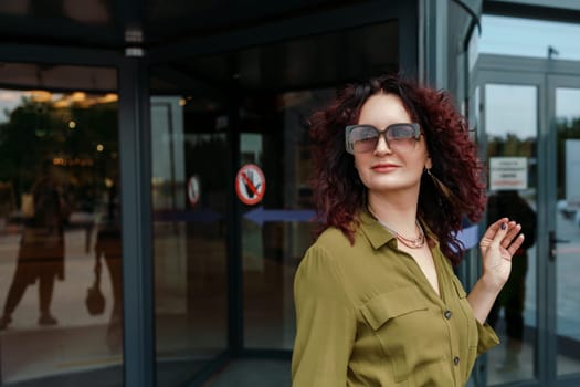 Woman leaves a supermarket. Caucasian model with long brunette hair, wears sunglasses and a khaki dress.