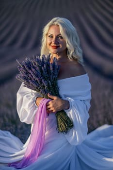 Blonde woman poses in lavender field at sunset. Happy woman in white dress holds lavender bouquet. Aromatherapy concept, lavender oil, photo session in lavender.