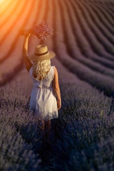 Blonde woman poses in lavender field at sunset. Happy woman in white dress holds lavender bouquet. Aromatherapy concept, lavender oil, photo session in lavender.