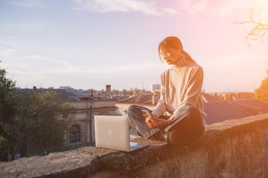 Woman freelancer uses laptop on cement wall outdoors against the sky and the roof of the city. The woman to be focused on her work or enjoying some leisure time while using her laptop