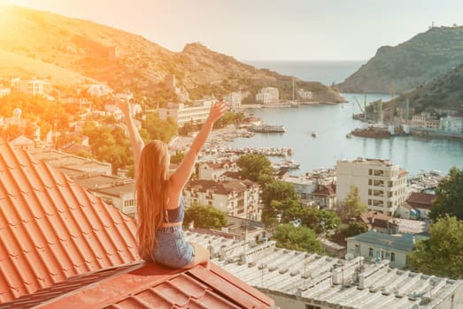 Woman sits on rooftop with outstretched arms, enjoys town view and sea mountains. Peaceful rooftop relaxation. Below her, there is a town with several boats visible in the water.