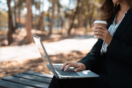 Young woman in formal suit drinking coffee and using laptop during working remotely in park.