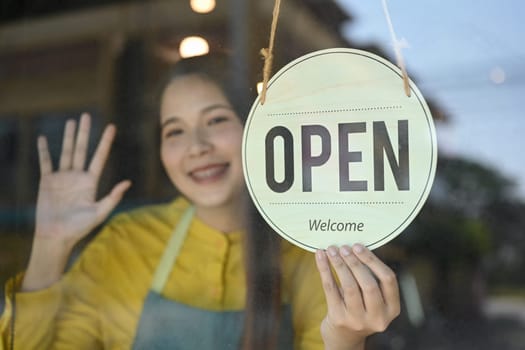 Smiling female small business owner wearing apron turning open sign on the glass door.