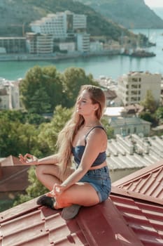 Woman sits on rooftop, enjoys town view and sea mountains. Peaceful rooftop relaxation. Below her, there is a town with several boats visible in the water. Rooftop vantage point