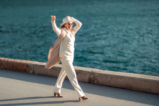 Happy blonde woman in a white suit and hat posing at the camera against the backdrop of the sea.