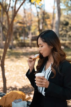 Beautiful young businesswoman with cup of coffee eating tasty sandwiches on park bench.